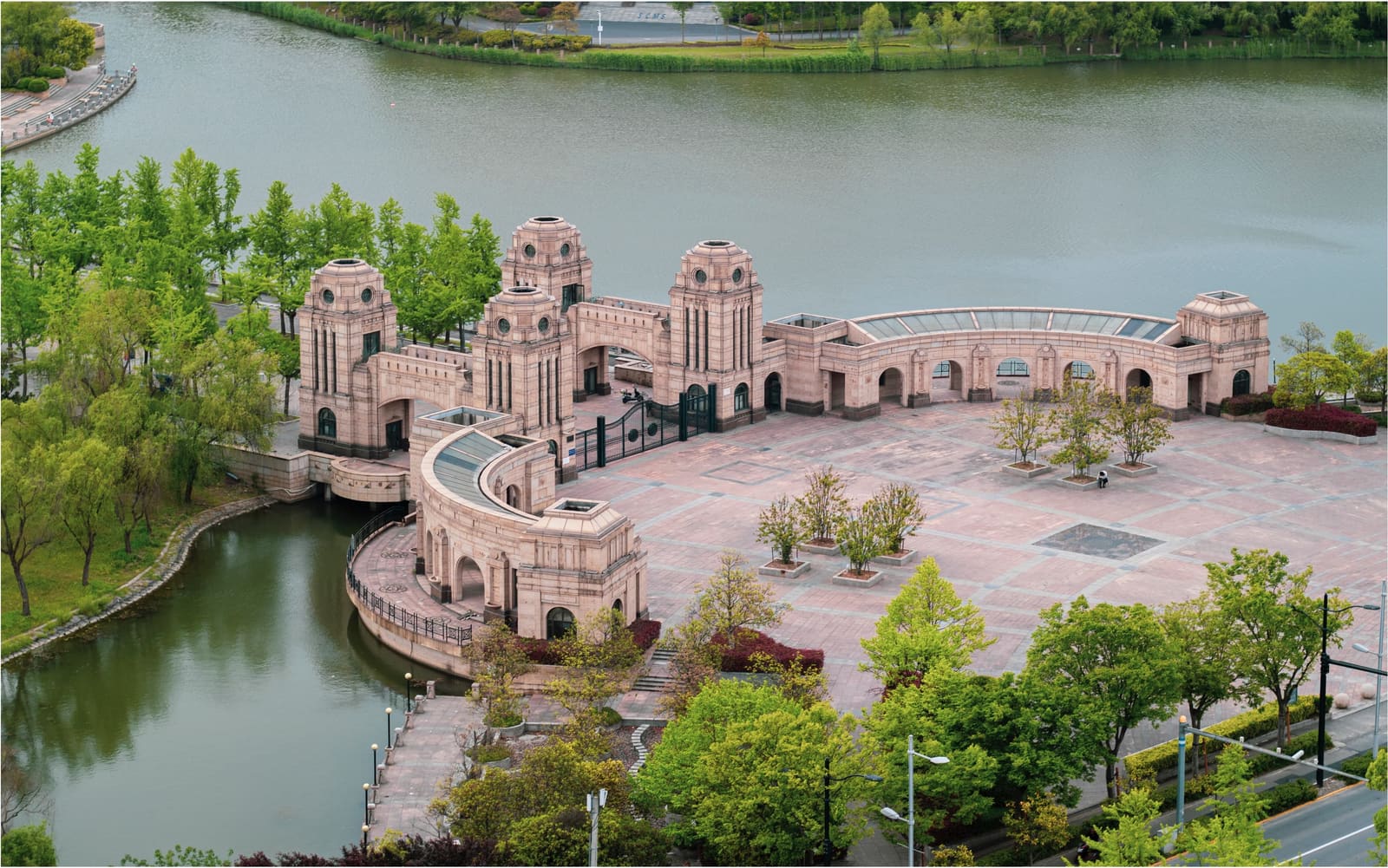 Aerial view of a sandstone gate complex with towers and curved arcades beside a lake on a Fudan University campus